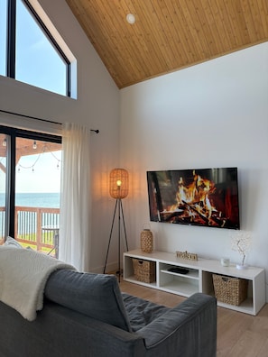 Ein modernes Wohnzimmer mit groĂem Fernseher, Holzboden, einem Balkon mit Blick und einer gemĂŒtlichen Couch.