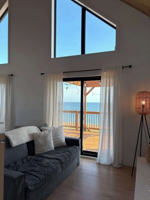Una habitación con un sofá gris oscuro, cortinas blancas, piso de madera y vista al mar a través de una puerta de cristal.