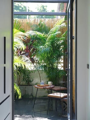 A glass door leading to a patio with a small table and chairs, surrounded by lush green plants and a wooden fence. - 10 min BTS 1Gbs WIFI NFLIX Full Kitchen HomeCinema (Khet Phra Khanong)