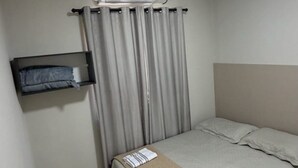 Quarto pequeno e bem arrumado, com cama, mesa de cabeceira, aquecedor fixado na parede e janela com cortinas.