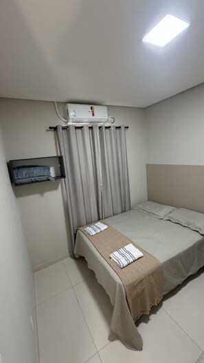 Un dormitorio pequeño y ordenado con cama, mesita de noche, calentador de pared y ventana con cortinas.