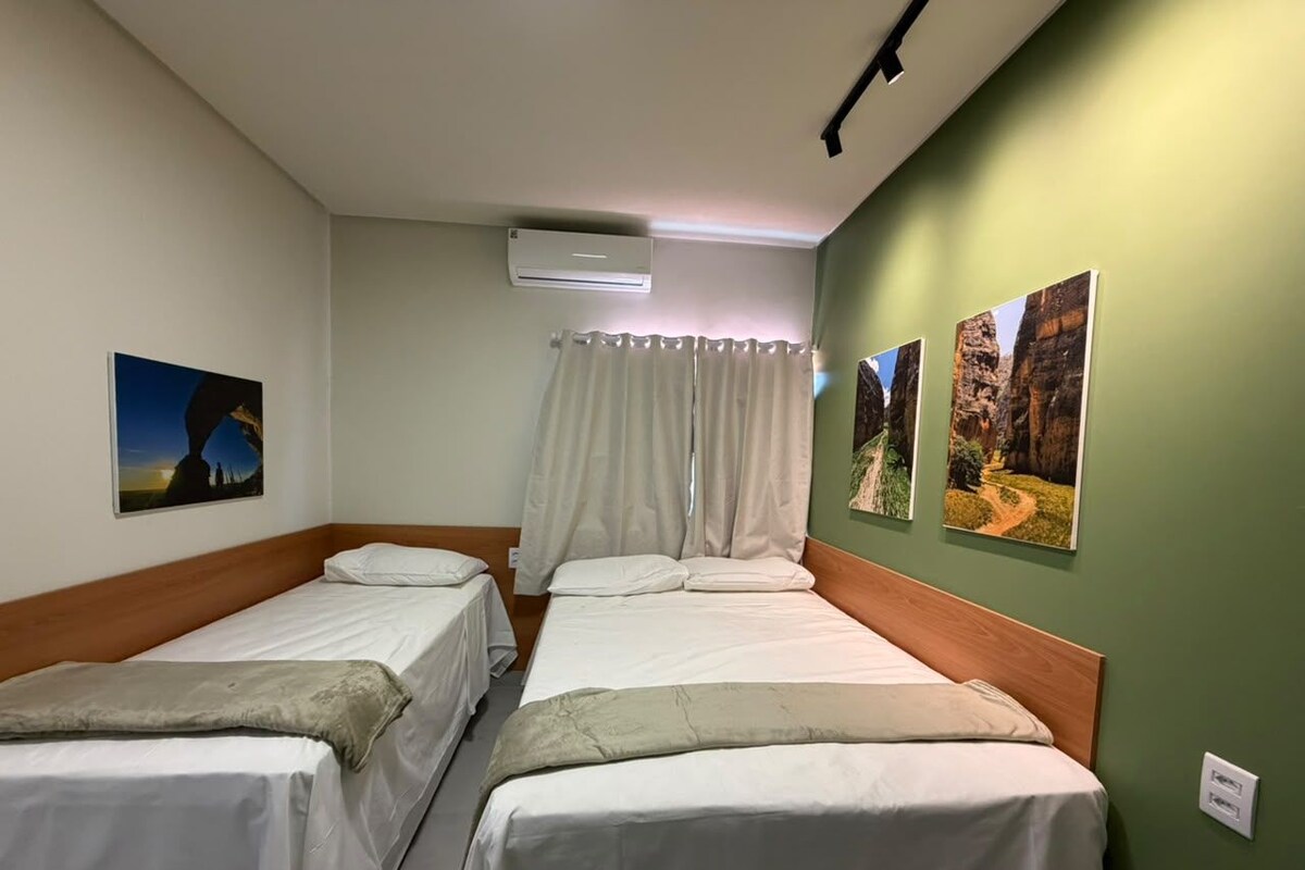 A hotel room with two single beds, a window with curtains, an air conditioning unit, and wall-mounted artwork.