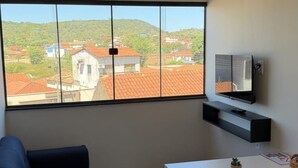 Uma sala de estar moderna com um sofá, uma mesinha, uma TV fixada na parede e uma grande janela com vista para edifícios e vegetação.
