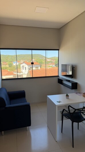 Una sala de estar moderna con un sofá, una mesita, un televisor montado en la pared y un ventanal con vistas a edificios y follaje.