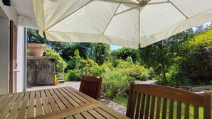 A wooden table and chairs under a large umbrella in a garden area with a view of greenery and a building. - In the shade of a beech tree: An oasis of tranquility just a few minutes from the lake (Miasino)