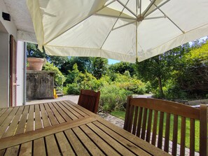 Ein Holztisch mit Stühlen steht unter einem großen Sonnenschirm in einem Garten mit Blick auf Grünflächen und ein Gebäude.