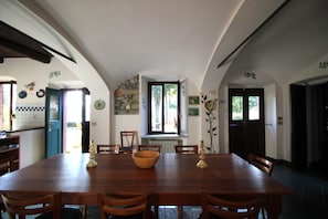 Ein Esszimmer mit einem großen Holztisch, Stühlen und einer Schüssel. Es gibt Fenster und Türen sowie eine Heizung an der Wand.