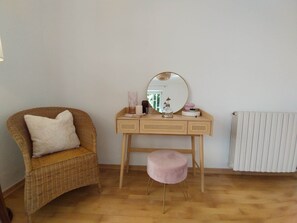 Ein Zimmer mit einem Korbstuhl, einem hölzernen Schminktisch mit Spiegel, einem pinken Hocker und einer Heizung.
