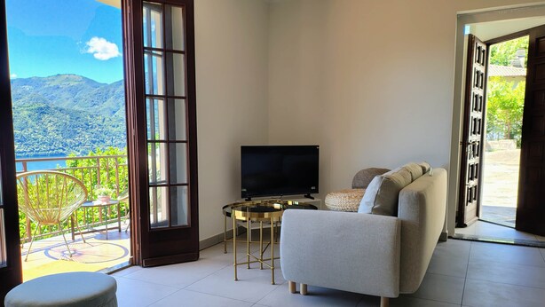 A living room with a sofa, a TV, a small table, and a balcony with a view of mountains. - Orta Blu Lake House: Spectacular view of Lake Orta, great (Miasino)