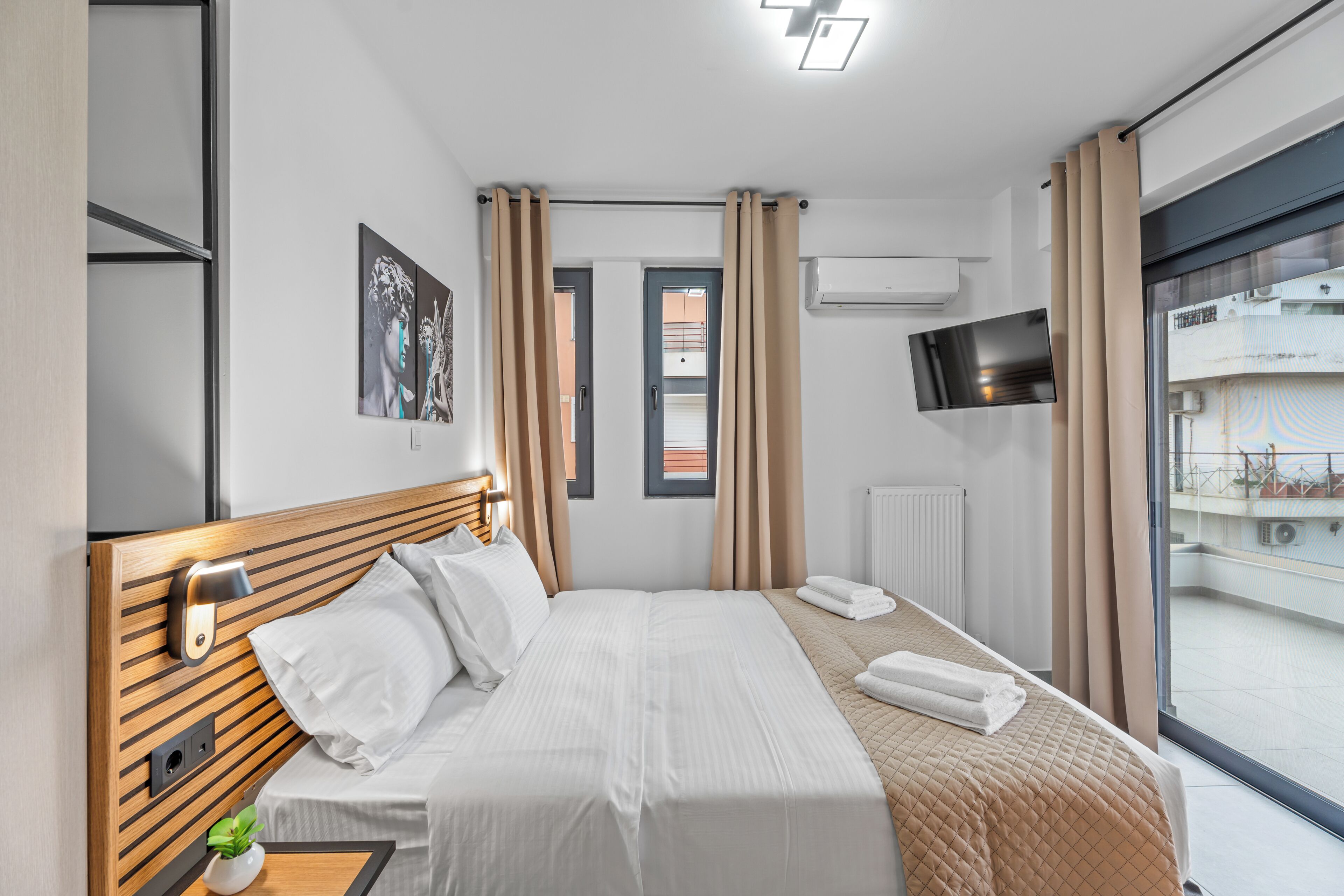 A modern hotel room with a wooden headboard, a flat-screen TV, a bed with white linens, a window with curtains, and a view of the city.
