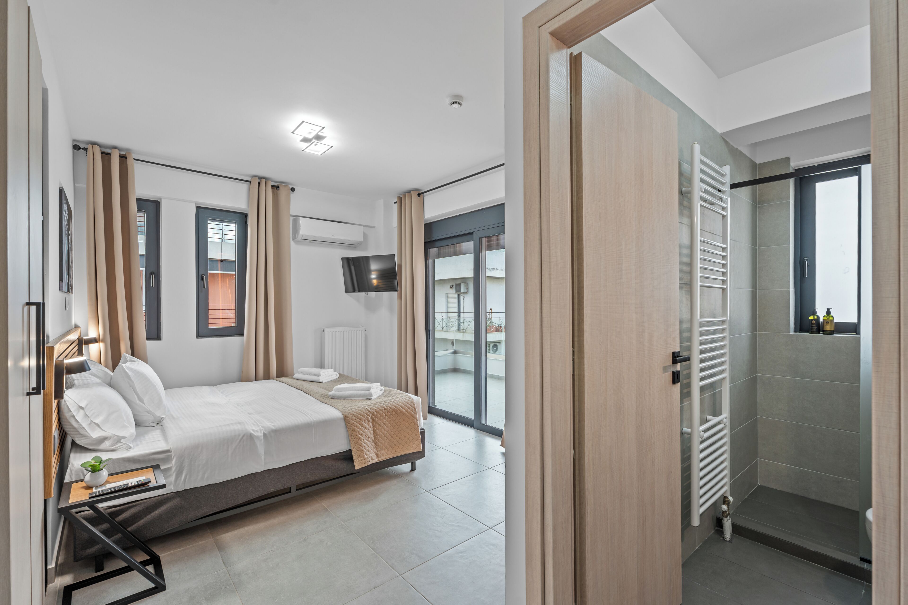 Een moderne slaapkamer met een bed, nachtkastje, televisie en uitzicht op een balkon.