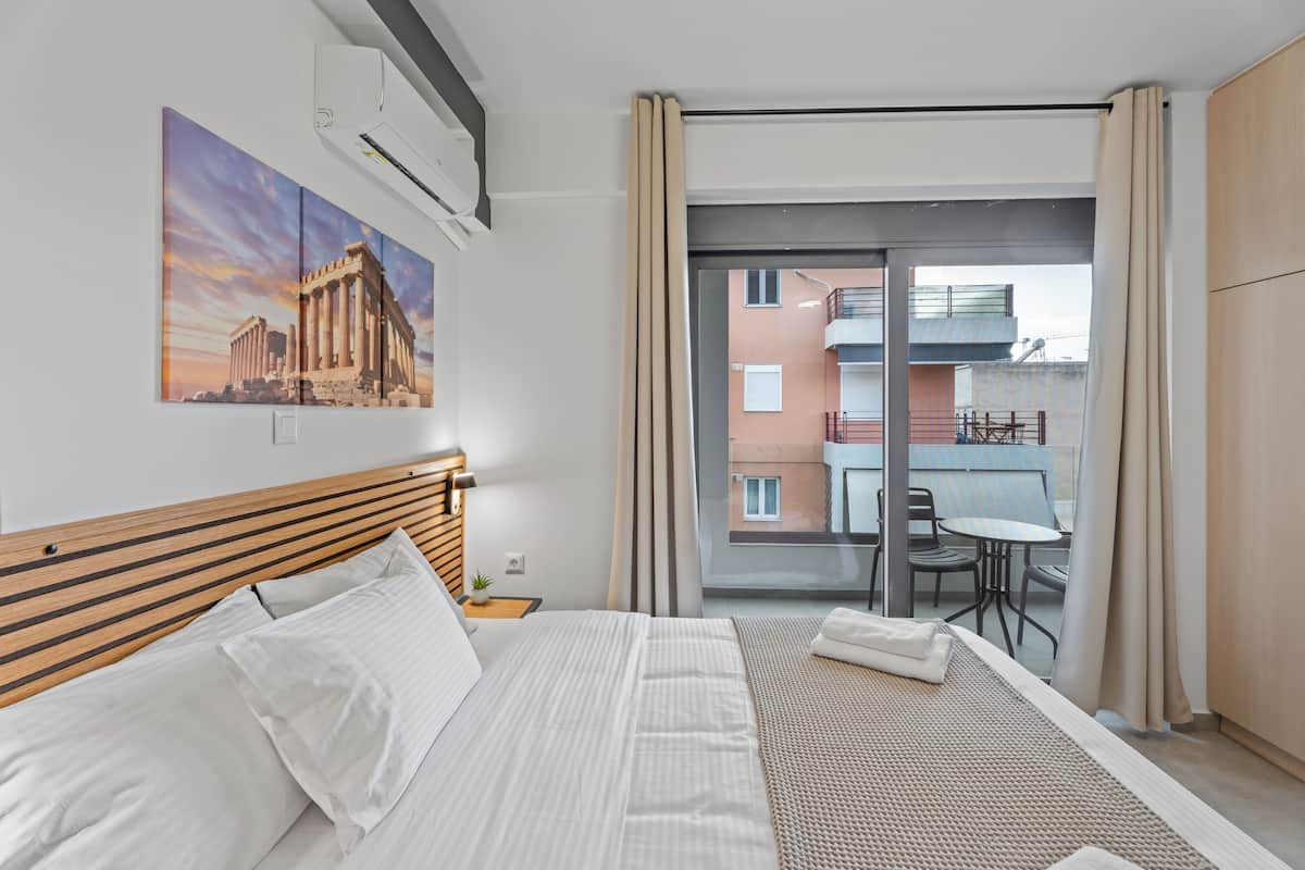 Quarto de hotel com cama, mesa de cabeceira, janela com cortinas, varanda com mesa e cadeiras, e um quadro de um edifício histórico.