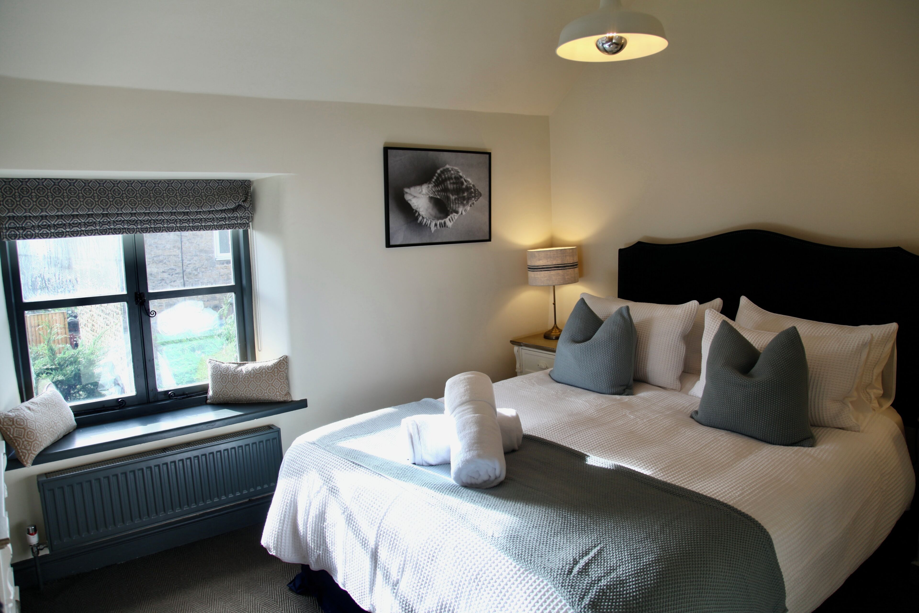 A bedroom with a bed, pillows, a window with a view, a radiator, a lamp, and a framed picture on the wall.