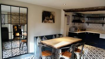 A dining area with a wooden table and metal chairs, a kitchen with shelves, and a stone wall.