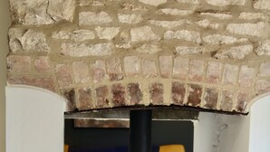 A stone fireplace with a black wood-burning stove, a sofa with patterned cushions, and a tiled floor. - Cosy Cotswold Cottage with Fireplace and Courtyard (Oxfordshire)