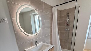 A modern bathroom with a wooden vanity, a round mirror, a shower with a glass door, and a tiled floor.