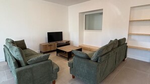A living room with two green sofas, a wooden coffee table, a TV on a stand, and a window with blinds.