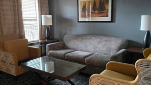 Habitación de hotel con un sofá gris, un sillón amarillo, una mesa de centro de vidrio, dos mesitas de noche con lámparas, un cuadro en la pared y una ventana con cortinas.