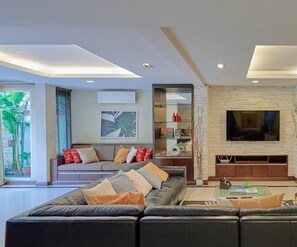 A modern living room with a large sectional sofa, a flat-screen TV mounted on a stone wall, and a glass coffee table. - Sleep10 | 100" Cinema | Quiet Gated Retreat | Mall (Khet Khlong Toei)
