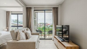A modern hotel room with a sofa, a TV on a wooden stand, and a balcony with outdoor furniture. - Comfy 1BR Getaway by Beach & Shops- diamond C176 (Tambon Choeng Thale)