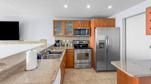 A modern kitchen with granite countertops, stainless steel appliances, wooden cabinets, and a flat-screen TV. - Hillside Village Hideaway | Sleeps 8 Comfortably (Mameyes II)