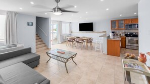 A modern living room with a sofa, coffee table, and dining area with chairs. There is a kitchen with appliances and a TV mounted on the wall. - Hillside Village Hideaway | Sleeps 8 Comfortably (Mameyes II)