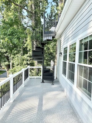Ein überdachter Außenbereich mit einer Wendeltreppe, die zu einem Balkon im zweiten Stock führt, umgeben von Bäumen und einem weißen Geländer.