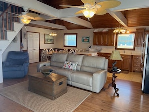 A spacious living area with a sofa, a coffee table, and a dining table with chairs. There is a staircase with a wooden railing leading to an upper level. The kitchen area is visible with wooden cabinets and a refrigerator.