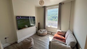 Ein Wohnzimmer mit einem Flachbildfernseher, einer Couch, einem Sitzsack und einem Fenster mit Blick auf Grünflächen.