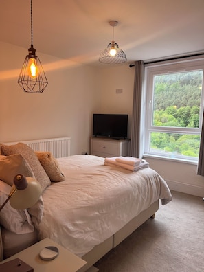 Une chambre à coucher comprenant un lit, une lampe de chevet, deux lampes suspendues, un téléviseur et une fenêtre donnant sur une forêt.