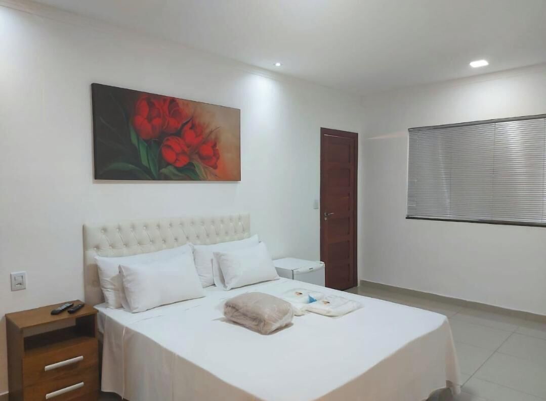 Un dormitorio con cama, mesita de noche, un cuadro, una puerta y una ventana con persianas.