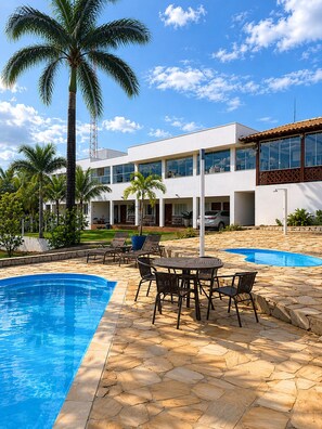 A modern building with a swimming pool, outdoor seating, and palm trees. - Suíte com Jacuzzi Vista de Escarpas 3 (Capitolio)