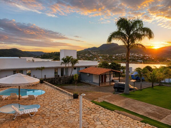 A pool area with lounge chairs, a stone patio, and a view of the sunset over a mountainous landscape.