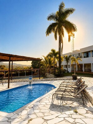 Una piscina con una cascada, rodeada de sillas de descanso y palmeras, con vista a un edificio al fondo.