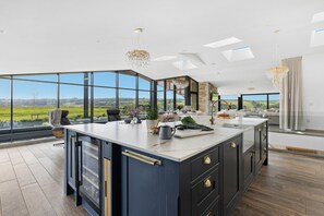 A modern kitchen with a large island, large windows, and a view of the countryside. - The Retreat at Strashleigh Manor Farm (Ivybridge)