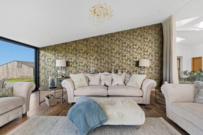 Un salon moderne avec un grand canapé, deux tables d’appoint, un lustre et une vue dégagée sur l’extérieur grâce à une grande fenêtre.