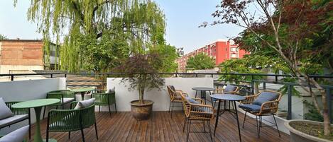 Una terraza de madera con mobiliario exterior, que incluye sillas y mesas, rodeada de árboles y un edificio al fondo.
