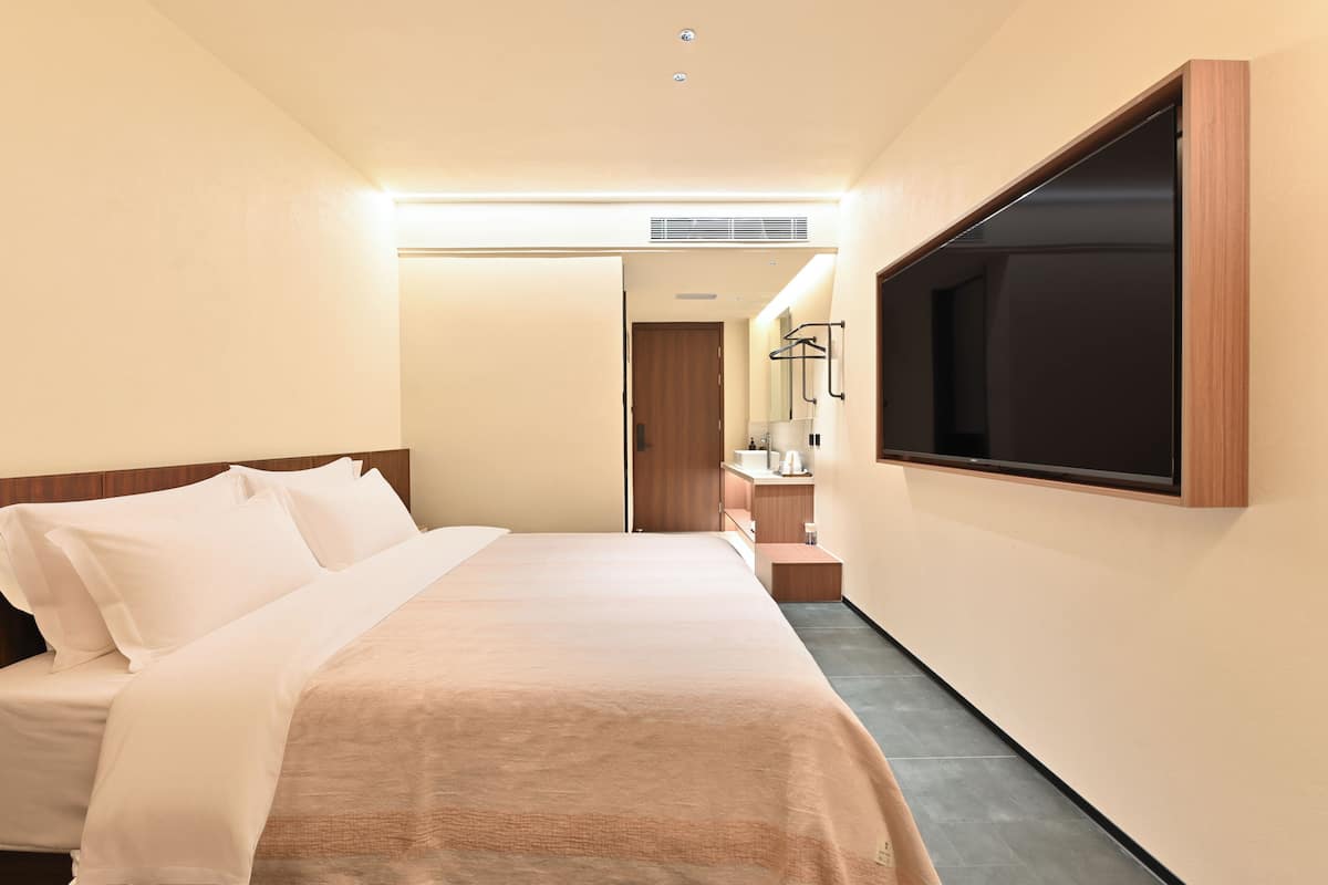 Una habitación de hotel con una cama, un televisor montado en la pared, un escritorio pequeño con un espejo y una puerta que da a un baño.