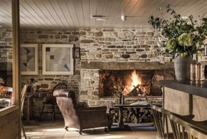 A stone fireplace with a fire, a wooden chair, and a table with a vase of flowers. - Cotswolds Grade II Listed Barn +underfloor heating (Little Rissington)