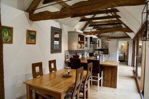 A dining area with a wooden table and chairs, a kitchen with white cabinets, and a loft bedroom with a sloped ceiling. - Cotswolds Grade II Listed Barn +underfloor heating (Little Rissington)