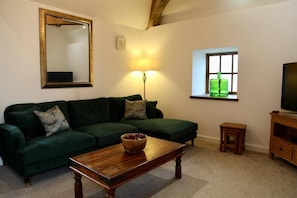 Een woonkamer met een groene bank, een houten salontafel, een vloerlamp, een televisie en een raam met uitzicht op een groene fles.