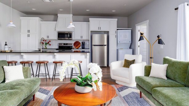 A modern kitchen with white cabinets, a microwave, and a refrigerator. There are two green sofas, a coffee table, and a bar stool area. - H3 Spacious & Urban Modern Condo - Great Location! (Oklahoma City)