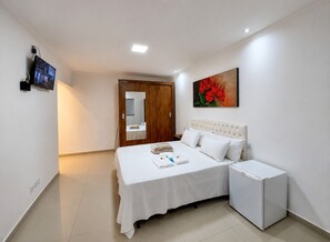 A bedroom with a bed, bedside table, a small refrigerator, a TV mounted on the wall, and a painting on the wall. - Suíte com Jacuzzi Vista de Escarpas 2 (Capitólio)