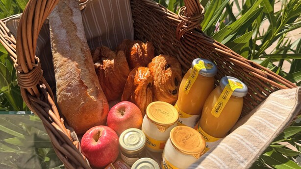 Ein Korb aus Weidwerk, gefüllt mit Lebensmitteln, darunter Brot, Gebäck, Äpfel und Saftflaschen, platziert im Freien vor einer Poolanlage und viel Grün.