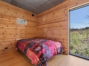 Ein Zimmer mit Holzwänden, einem Bett mit bunter Steppdecke, einem Fenster und Blick auf Grünfläche.
