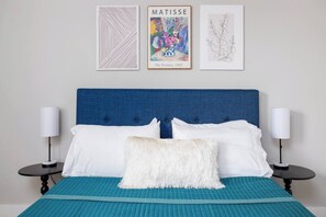 Ein Schlafzimmer mit einem blauen Kopfteil, weißen Kissen, einem flauschigen weißen Kissen und zwei Nachttischen mit Lampen.