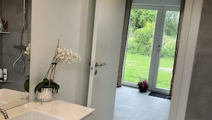 A bathroom with a white sink, a shower, and a door leading to a balcony with a view of greenery.