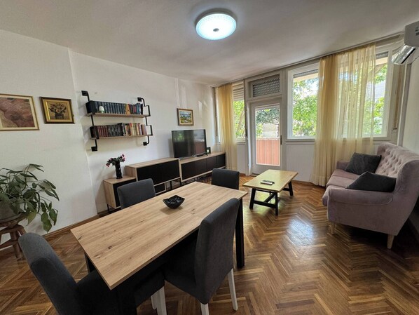 Ein Wohnzimmer mit Esstisch aus Holz, Stühlen, einer Couch, einem Fernseher und einem Bücherregal.