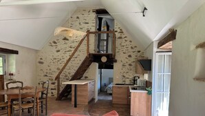 Un habitación estilo loft con techo alto, pared de piedra, escalera de madera, zona de cocina y mesa de comedor con sillas.