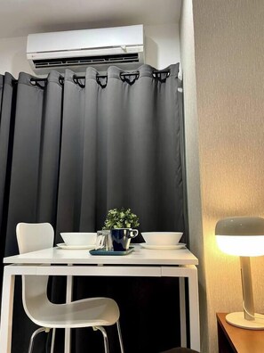 Ein Zimmer mit einem weißen Tisch, zwei Stühlen, einer kleinen Pflanze, einer Lampe und einem schwarzen Vorhang.
